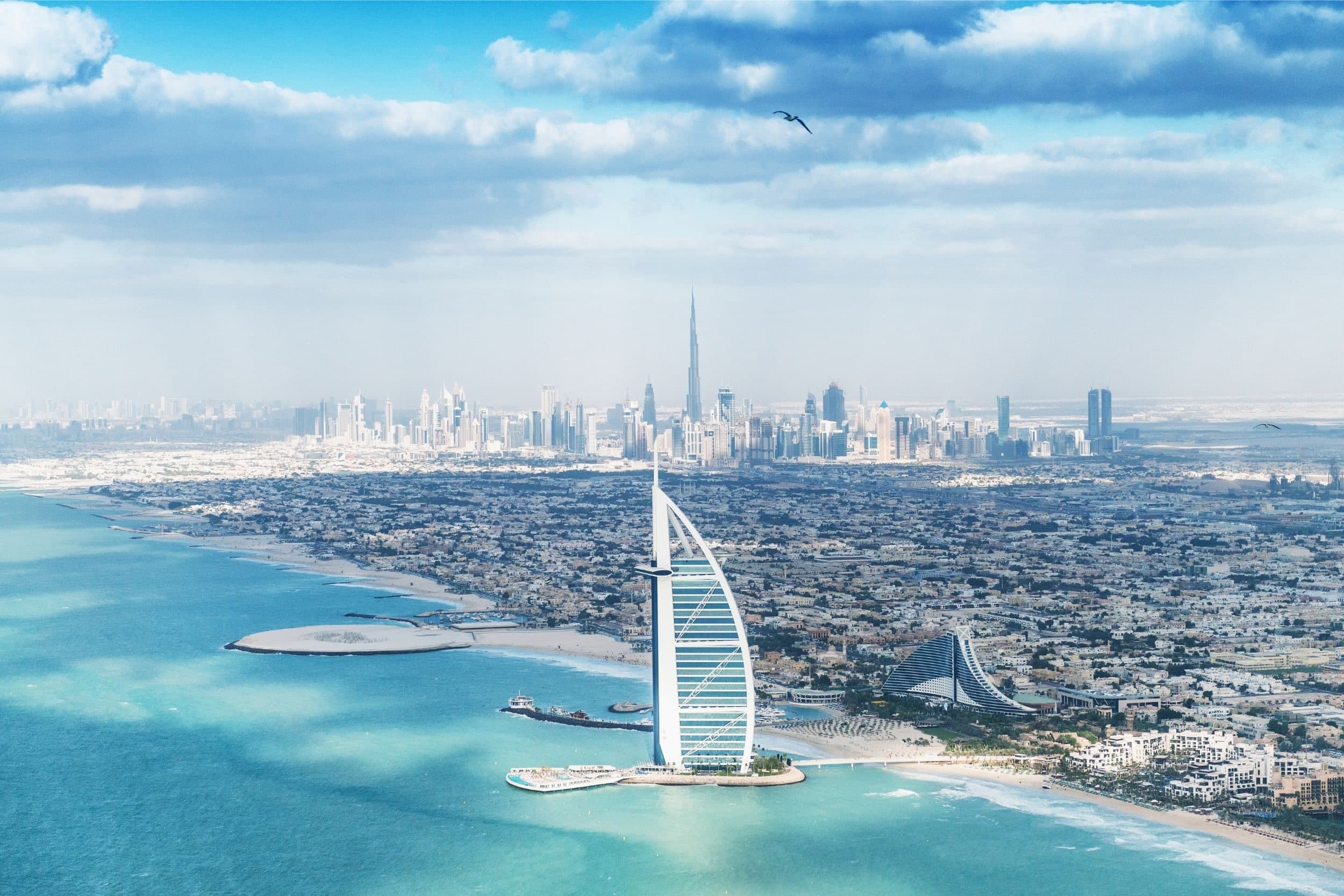 The Jumeirah coast from the air, with the Burj Al Arab and Jumeirah Beach Hotel in front, low-rise villa neighbourhoods along the beach and the Downtown Dubai skyline in the distance