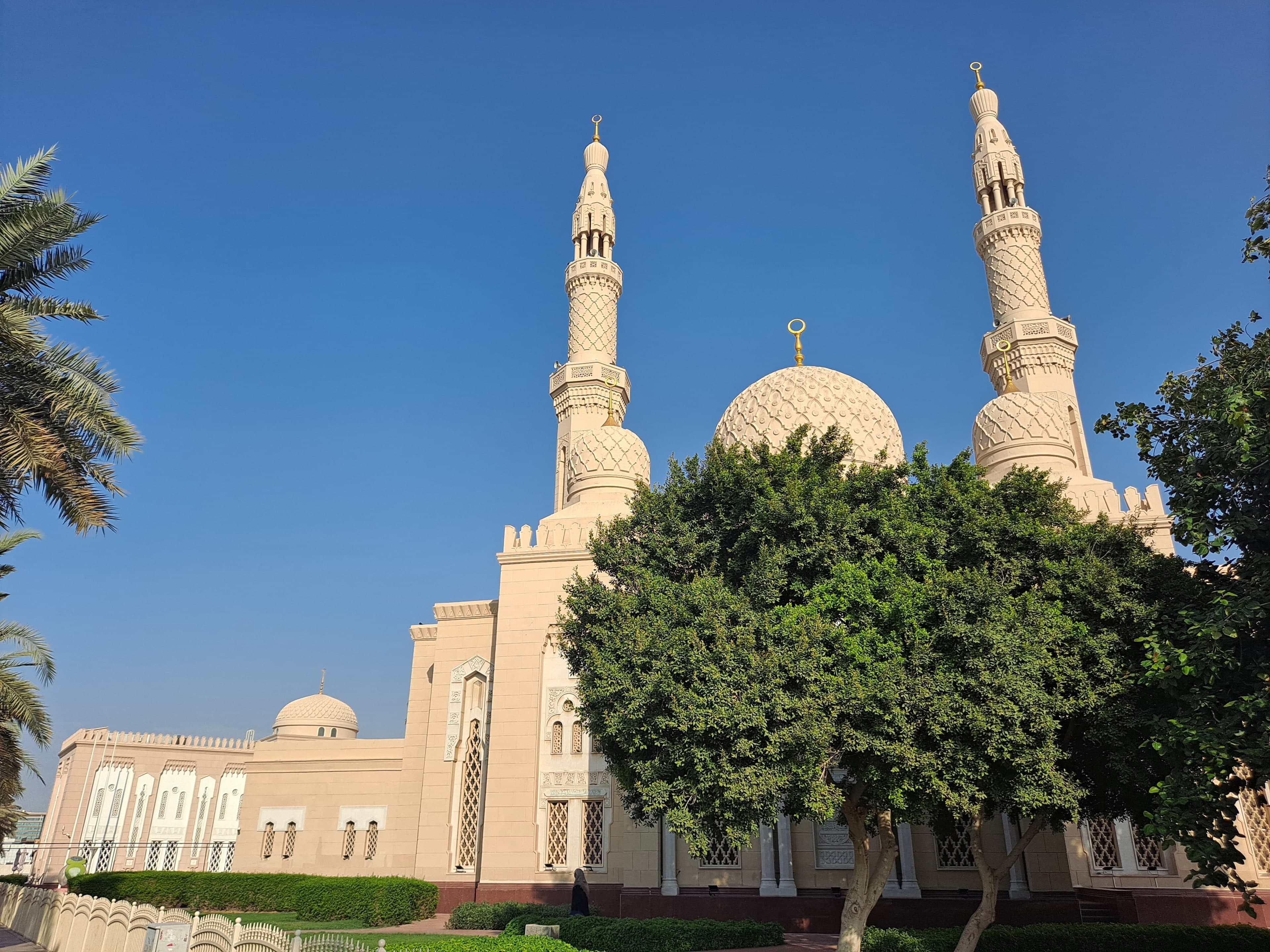 Jumeirah Mosque in pale stone with its two minarets and central dome, behind green trees under a blue sky
