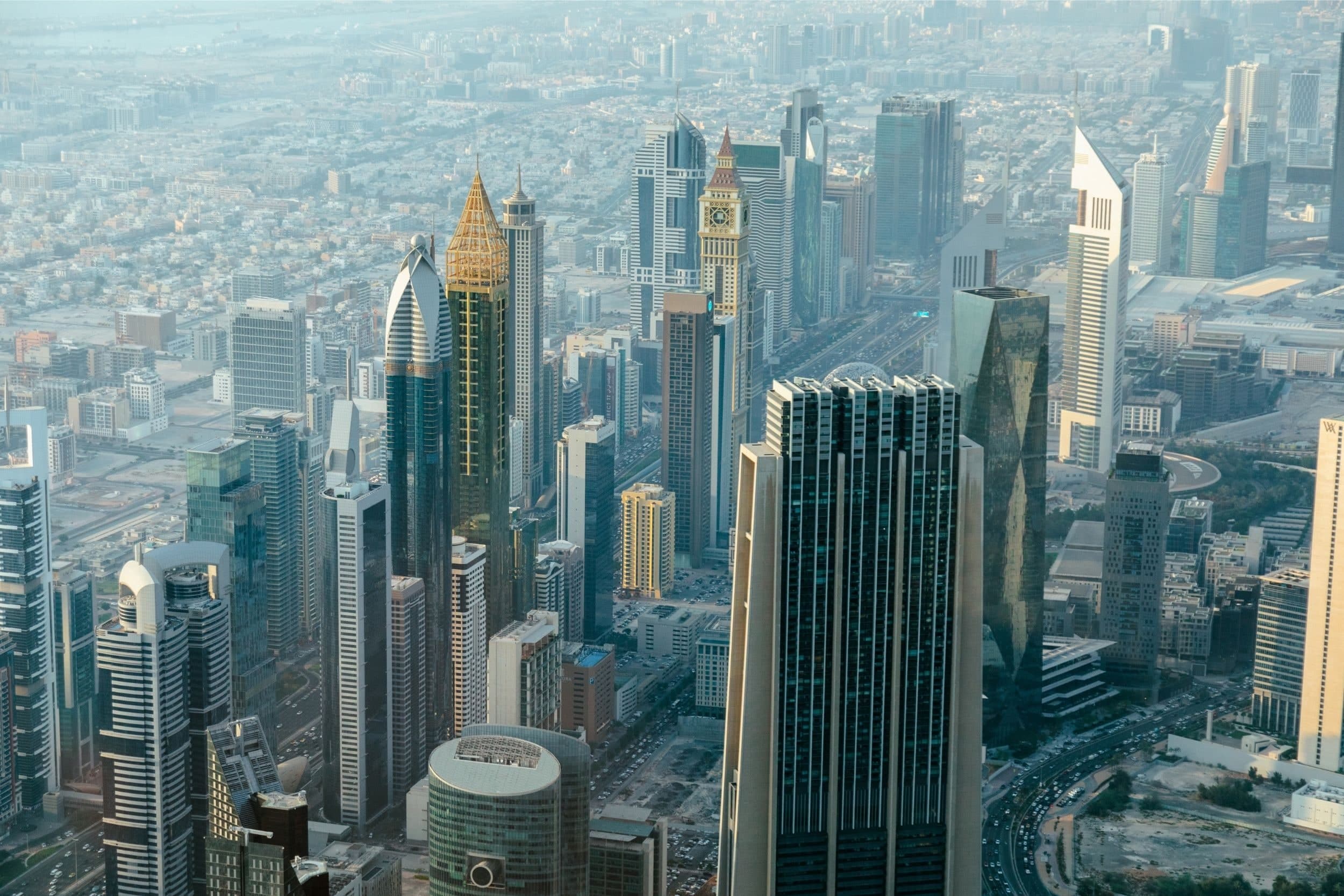The towers of DIFC and Sheikh Zayed Road seen from high above, with the Emirates Towers on the right and low-rise city stretching into the haze behind