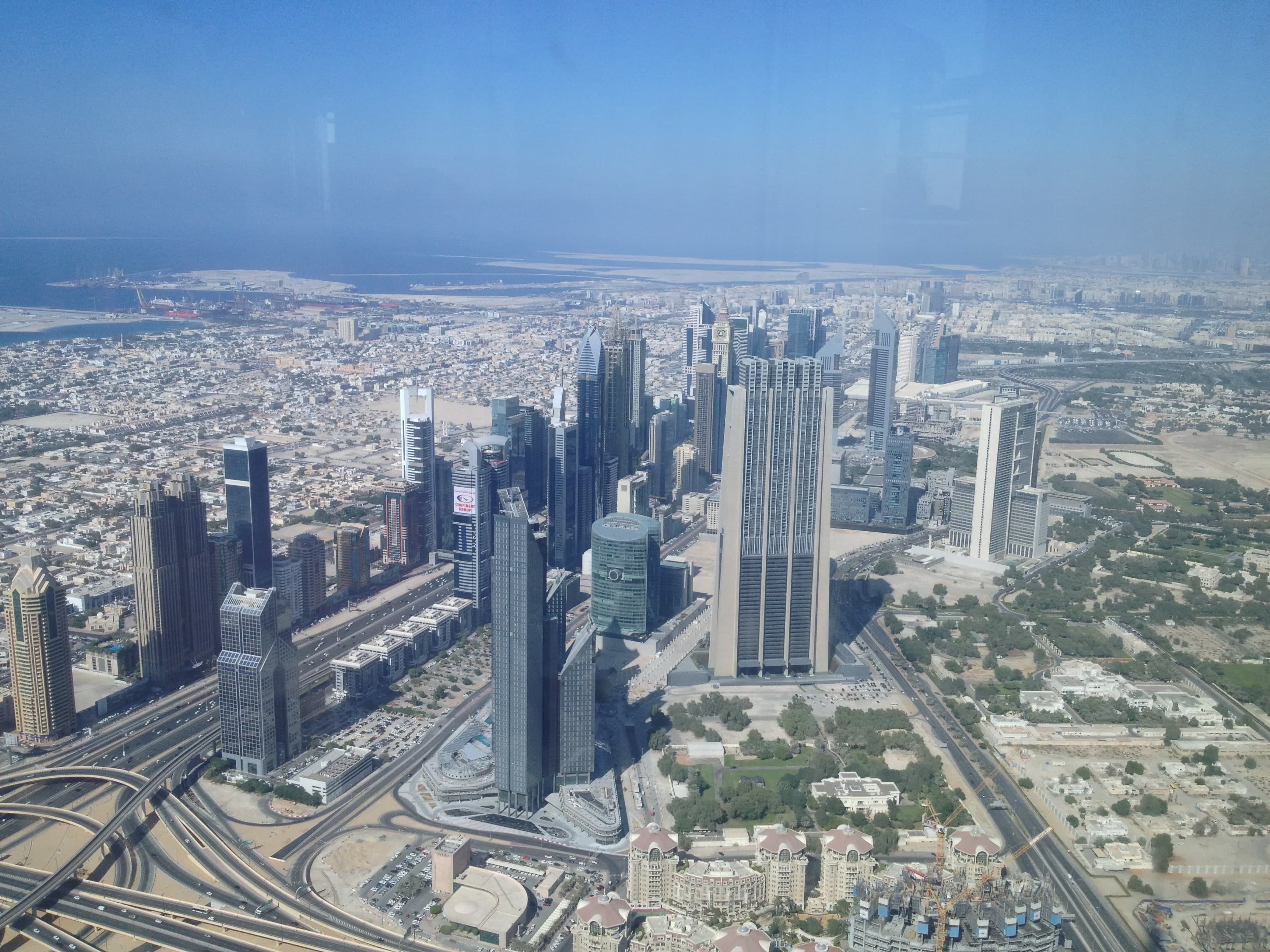 DIFC seen from the Burj Khalifa on a clear day, with its office and residential towers beside Sheikh Zayed Road and the sea in the distance