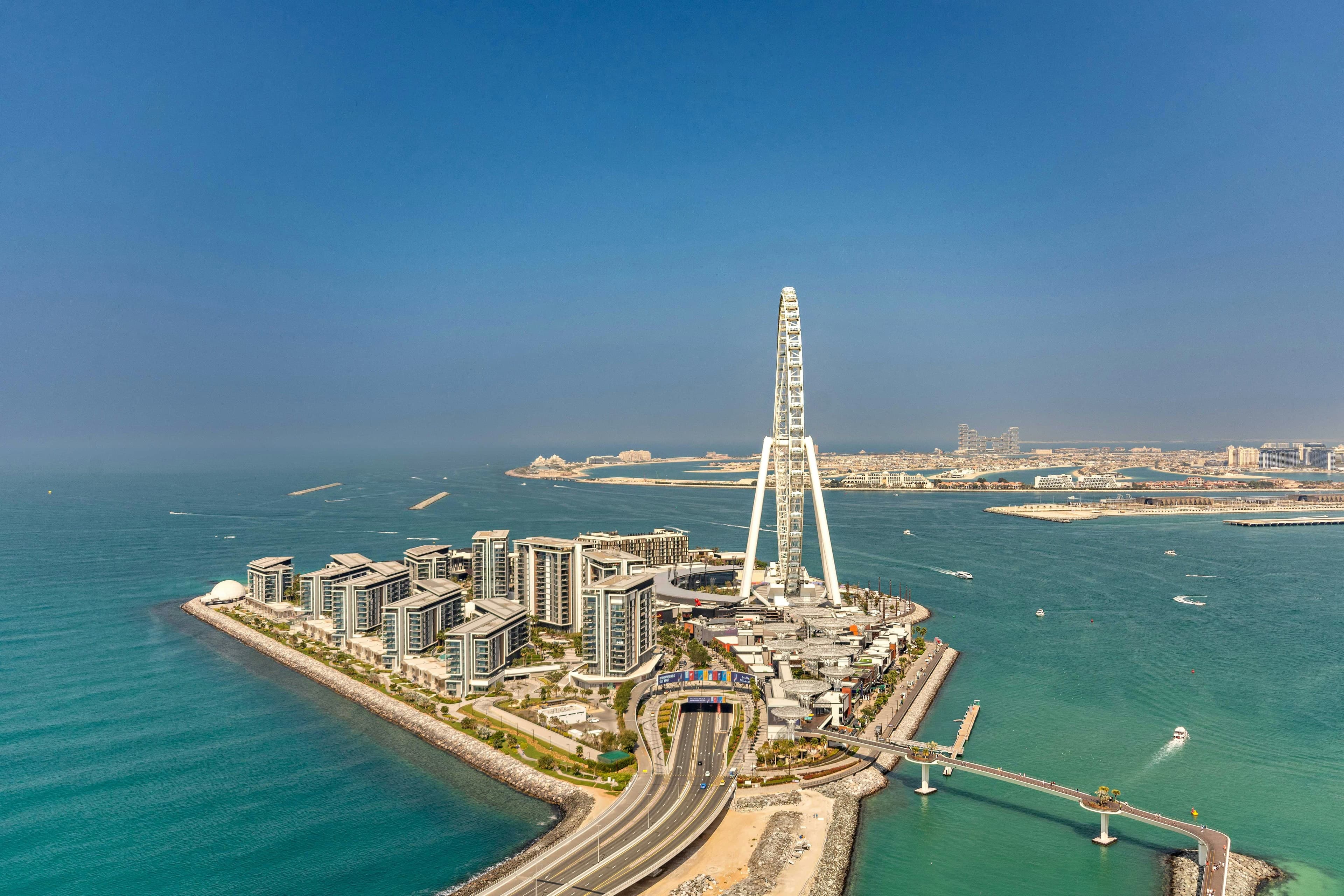 An aerial view of Bluewaters Island, with its low-rise residential buildings and the Ain Dubai observation wheel, and Palm Jumeirah on the horizon
