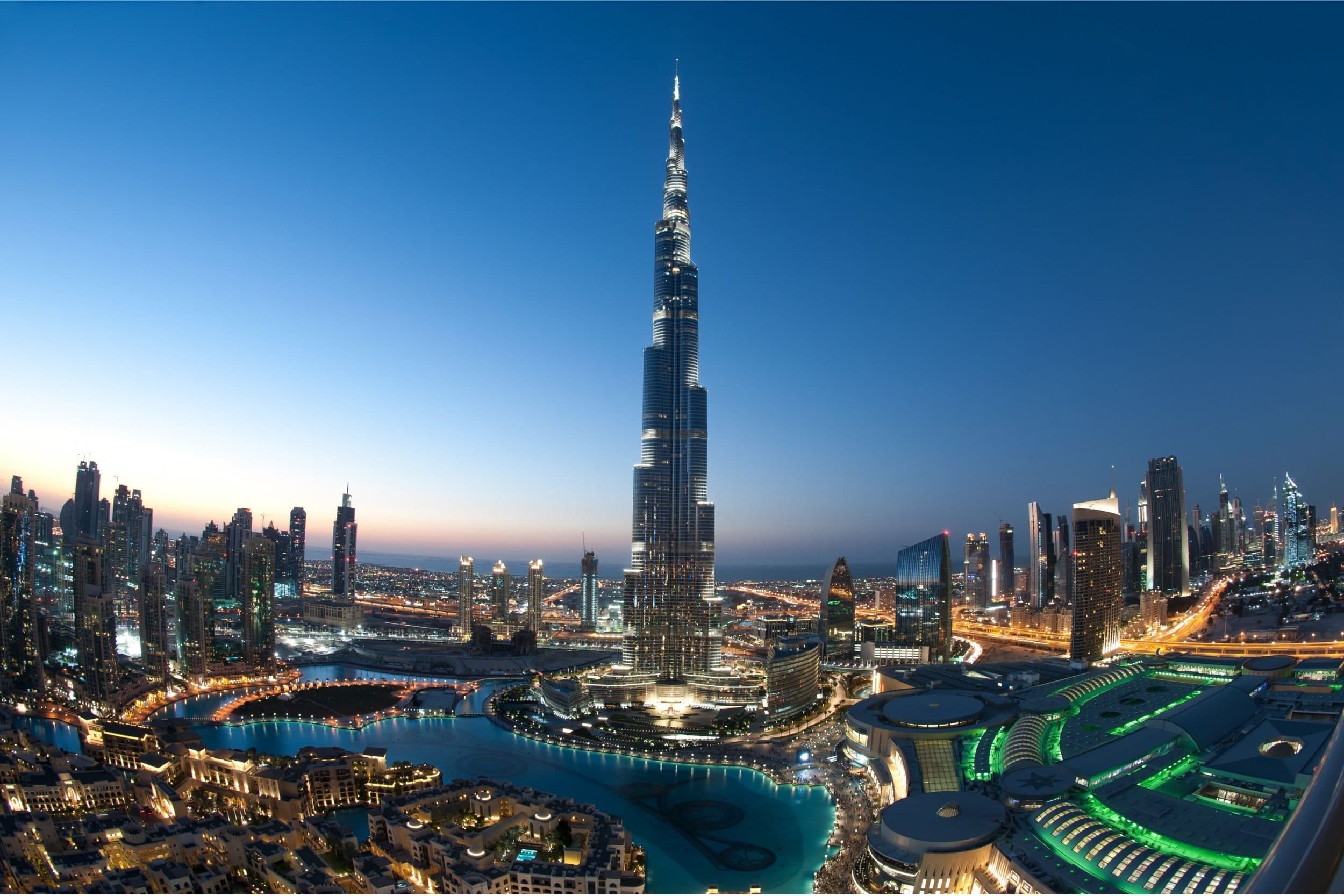 The Burj Khalifa at dusk seen from high above Downtown Dubai, with the Dubai Mall lit in green and the Burj Lake below