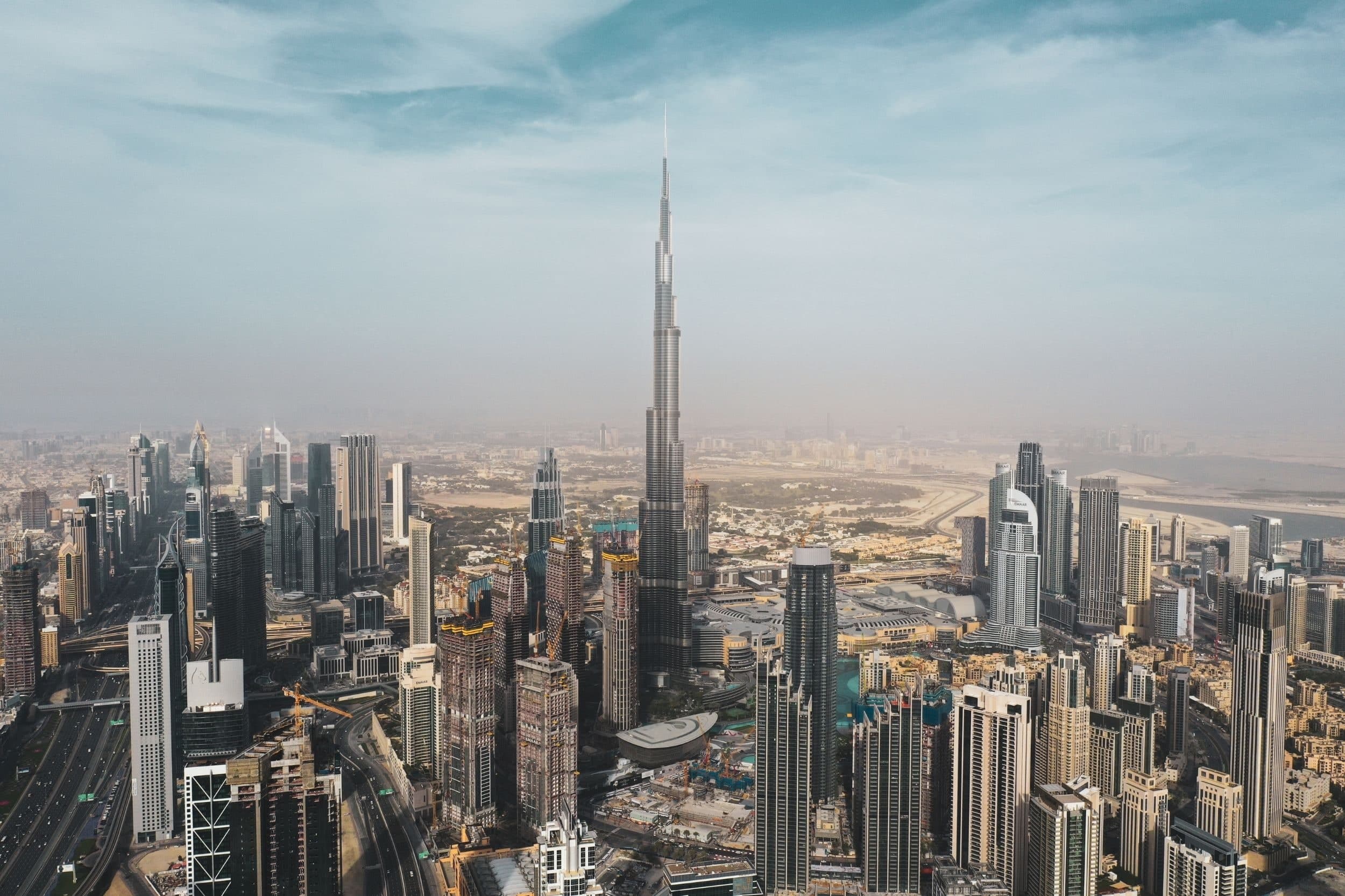 Downtown Dubai from the air on a hazy day, the Burj Khalifa rising above the surrounding towers, with a wide highway on the left