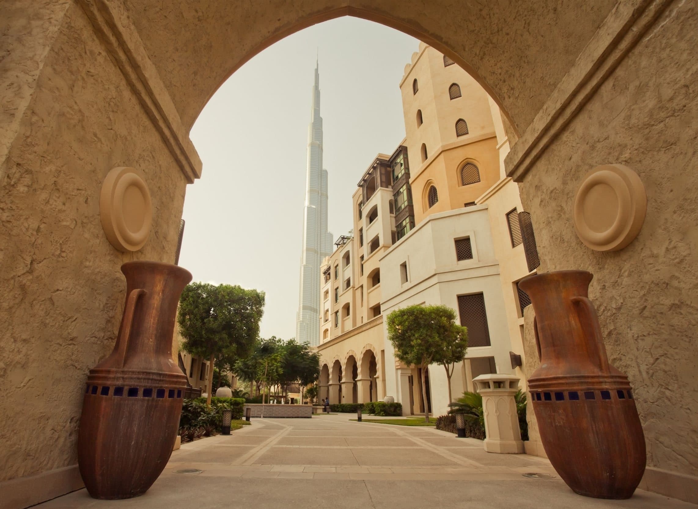 The Burj Khalifa framed by a stone archway in Old Town, with a paved walkway, trees and low-rise Arabic style buildings