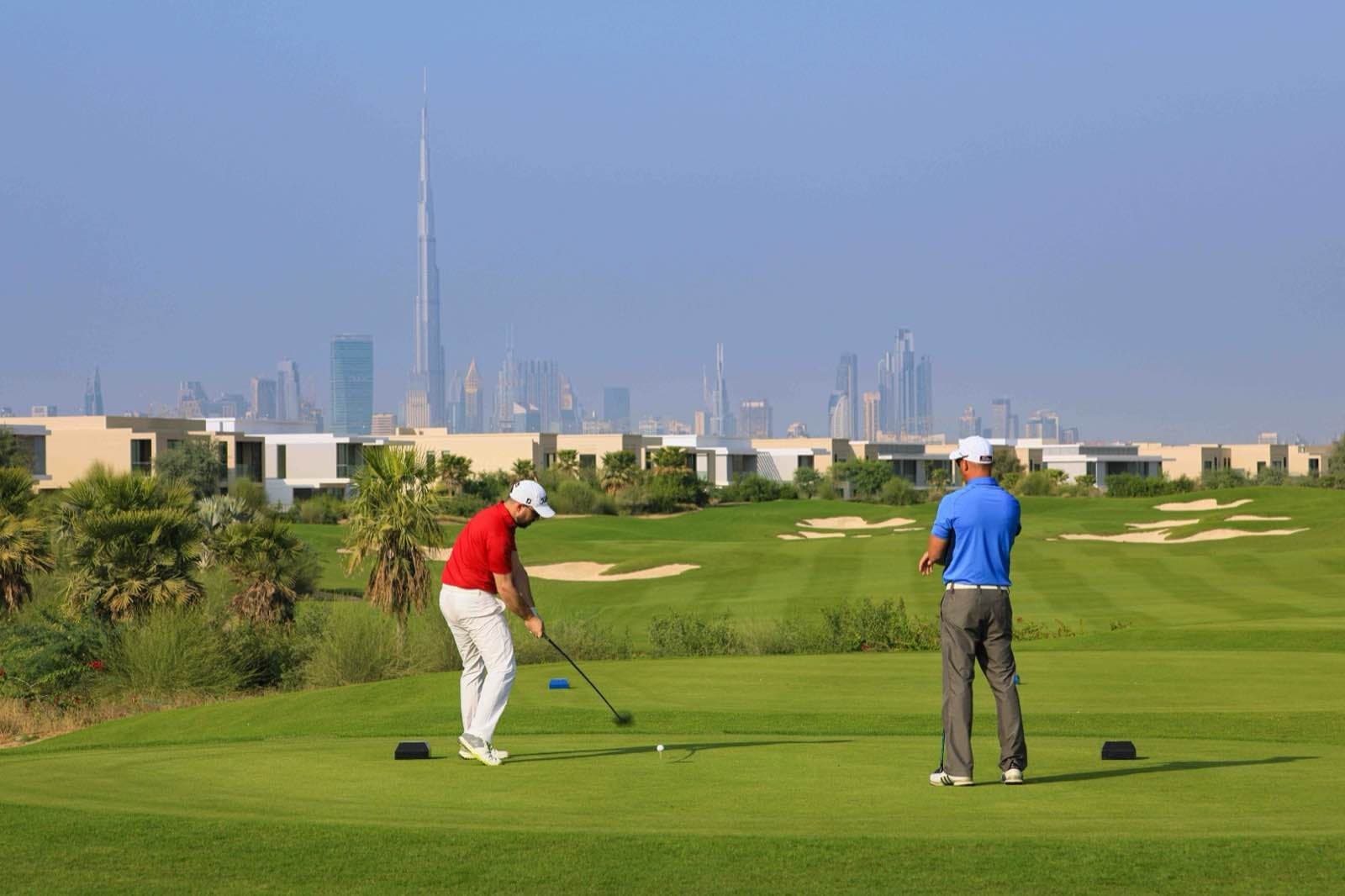 Two golfers on a tee at Dubai Hills Golf Club, with modern villas along the course and the Downtown Dubai skyline behind