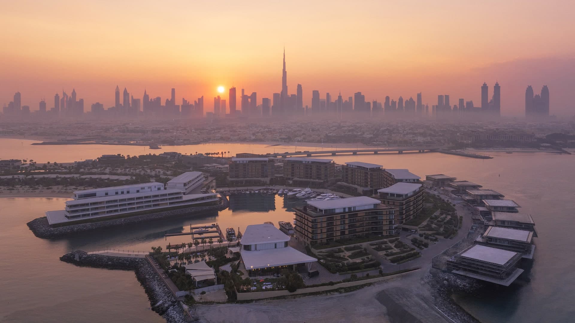 An aerial view of Jumeirah Bay Island at sunset, with the Bulgari Resort and Residences around the marina, the bridge to the mainland and the Downtown Dubai skyline behind
