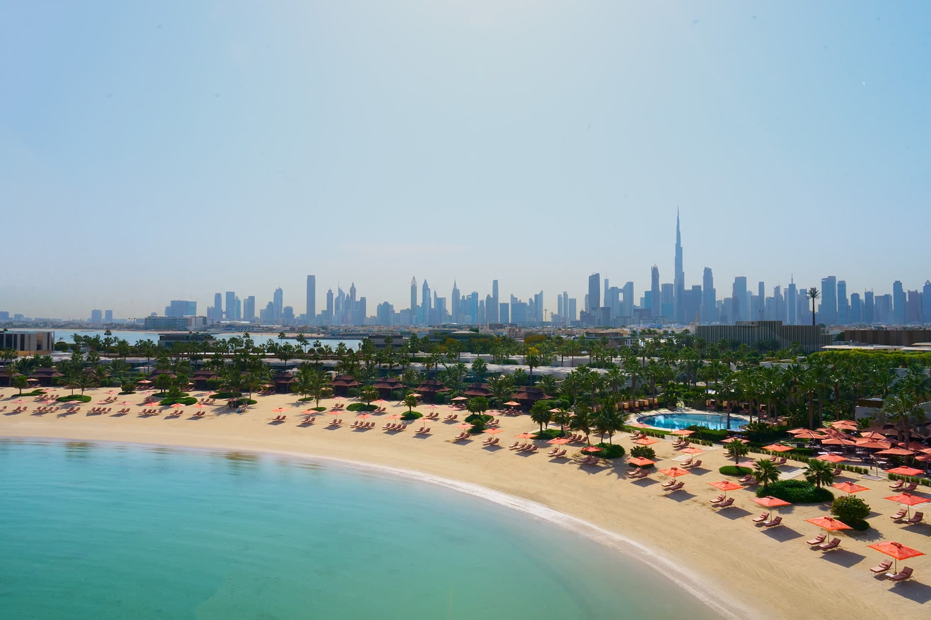 The curved private beach of the Bulgari Resort on Jumeirah Bay Island, lined with sun loungers and palm trees, with the Dubai skyline across the water