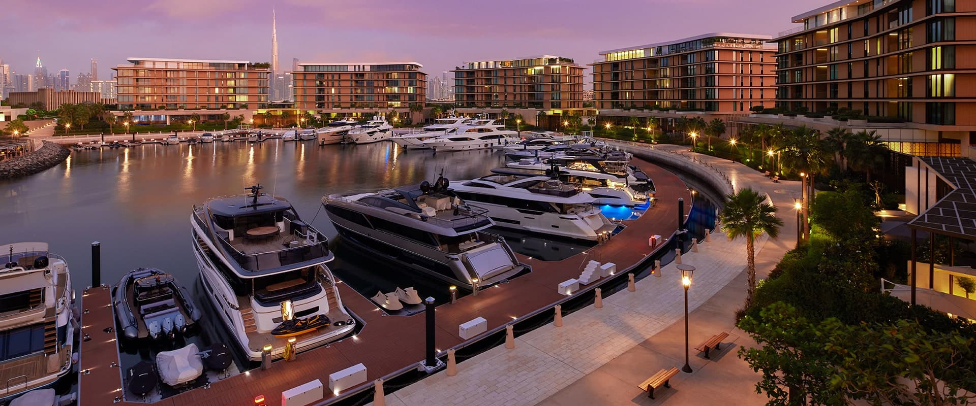 Yachts moored in the Bulgari marina on Jumeirah Bay Island at dusk, with the low-rise Bulgari Residences buildings around the water and the Burj Khalifa in the distance