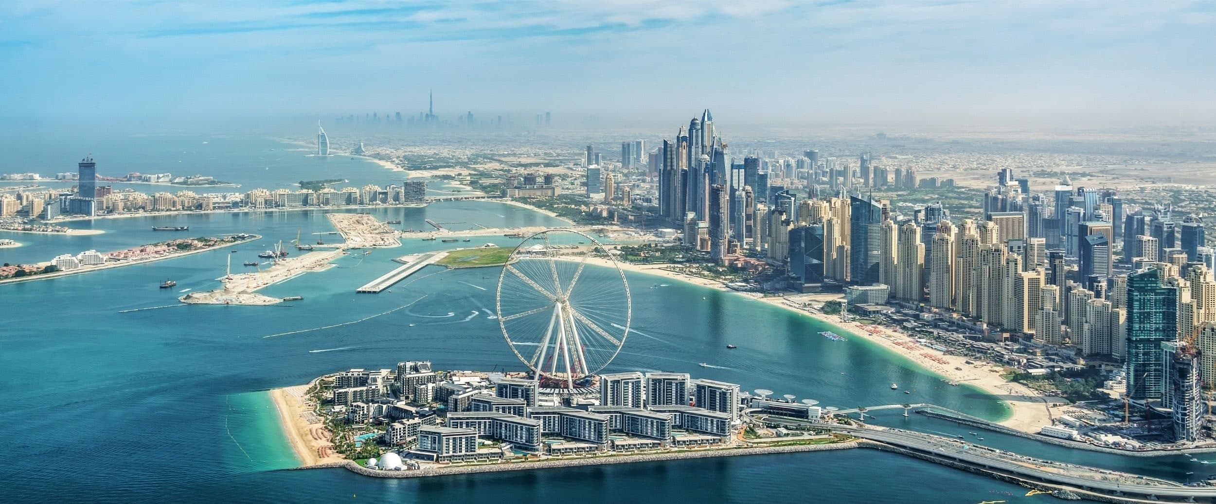 An aerial view of Bluewaters Island and its observation wheel, with the towers of JBR and Dubai Marina along the curving beach behind it