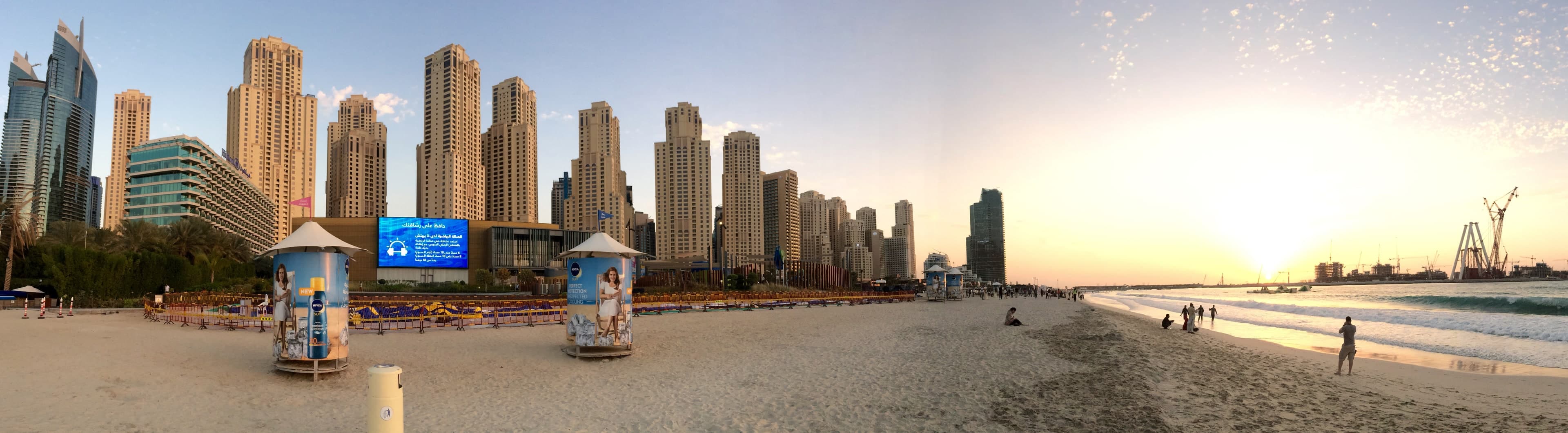 A wide view of the JBR towers rising behind the beach at sunset, with people walking along the water's edge
