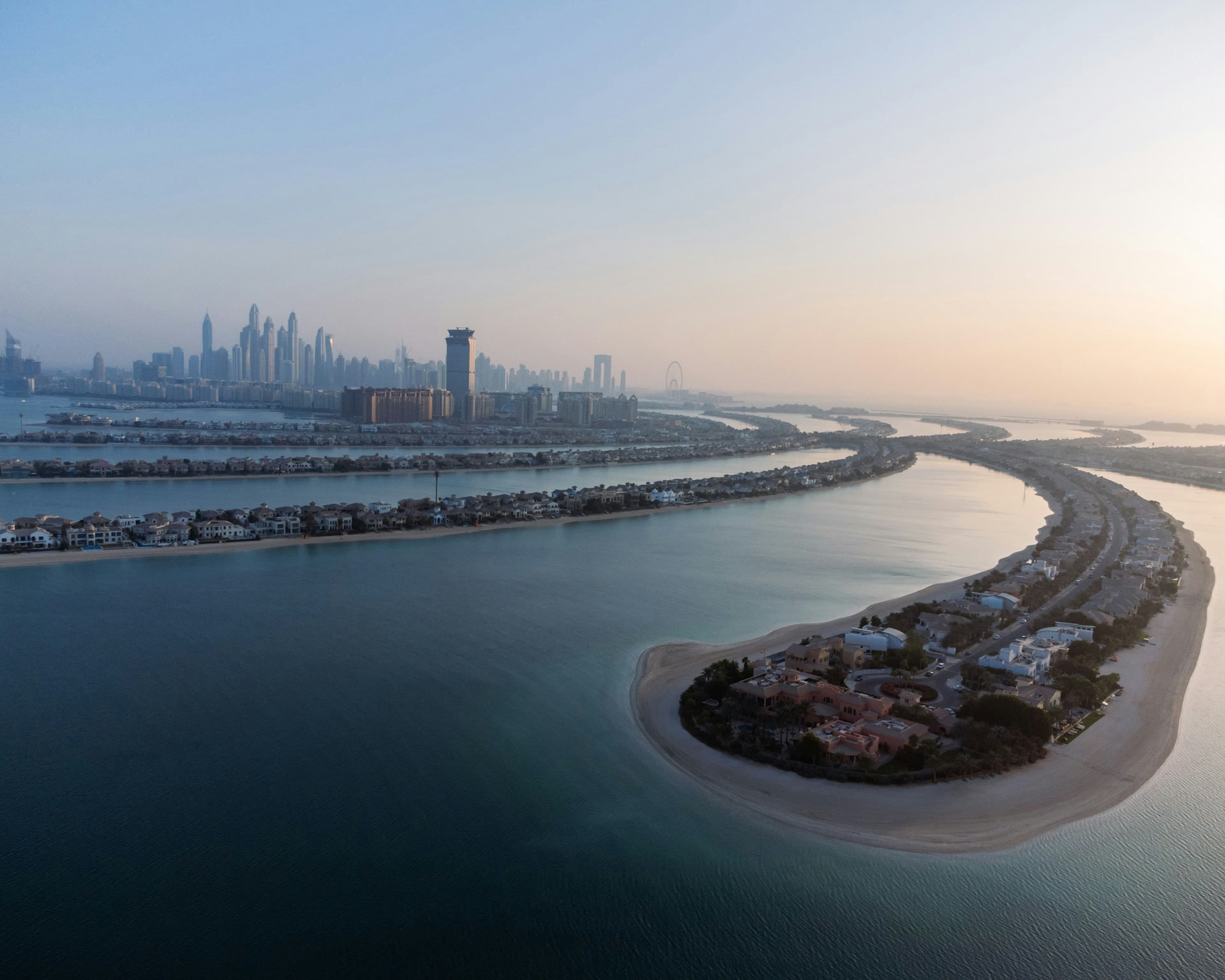 The tip of a Palm Jumeirah frond with villas and a sandy beach at dawn, and the Dubai Marina towers in the distance