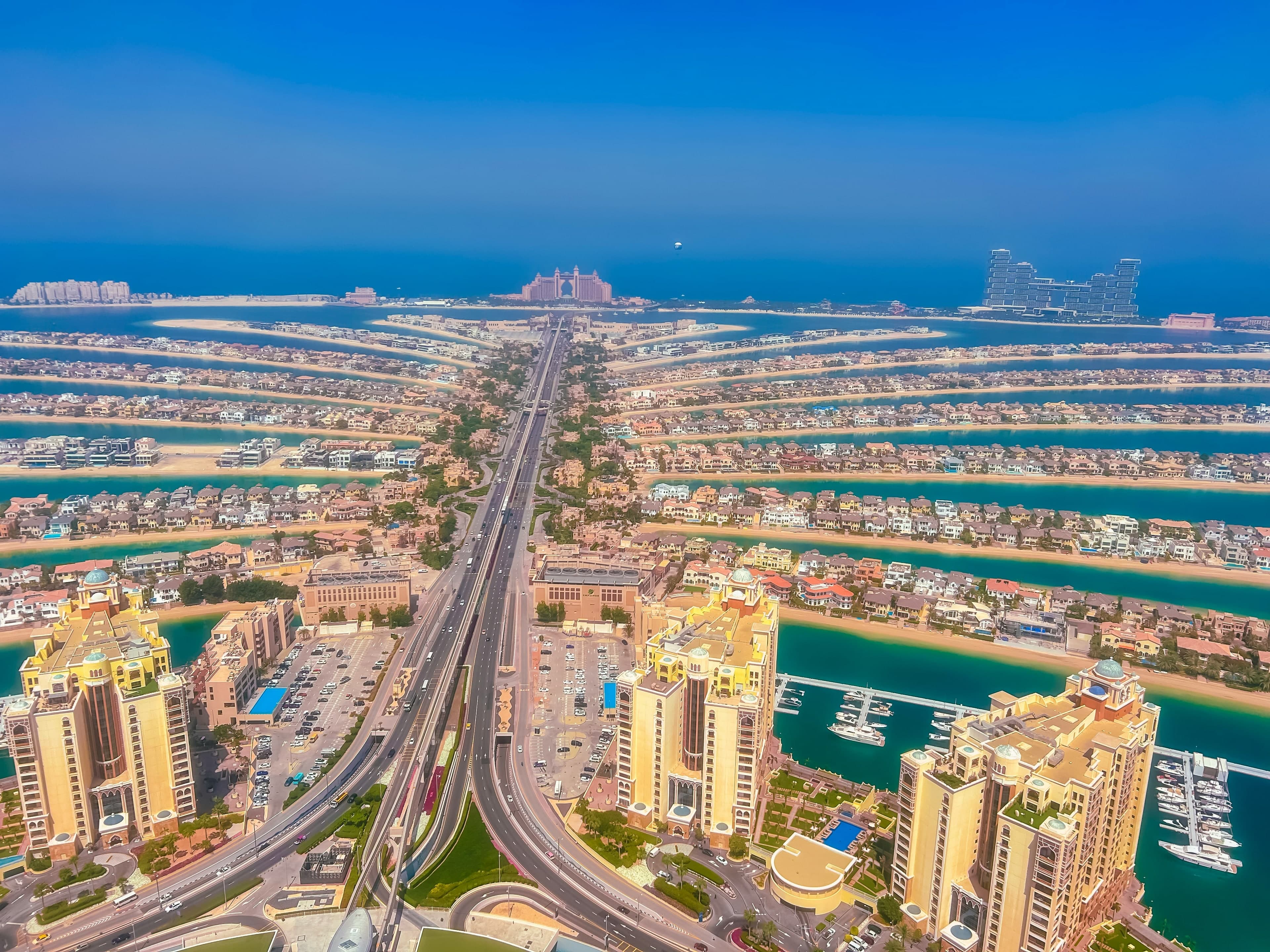 The fronds of Palm Jumeirah spreading out from the trunk on a sunny day, with apartment towers and a marina in the foreground and the crescent hotels behind