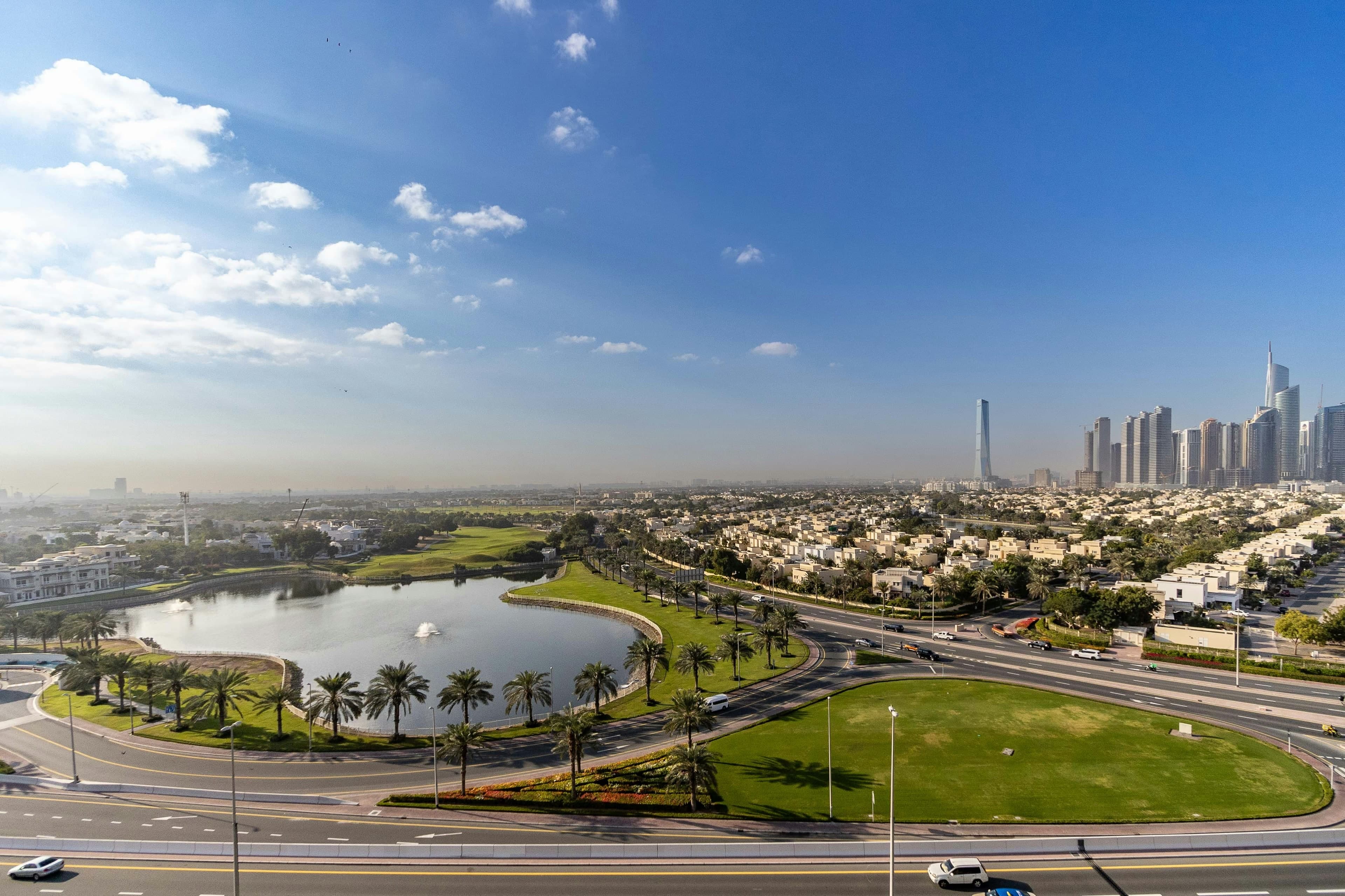 The Montgomerie golf course and lake in Emirates Hills seen from a high viewpoint, with villas around them and the towers of Jumeirah Lakes Towers on the horizon