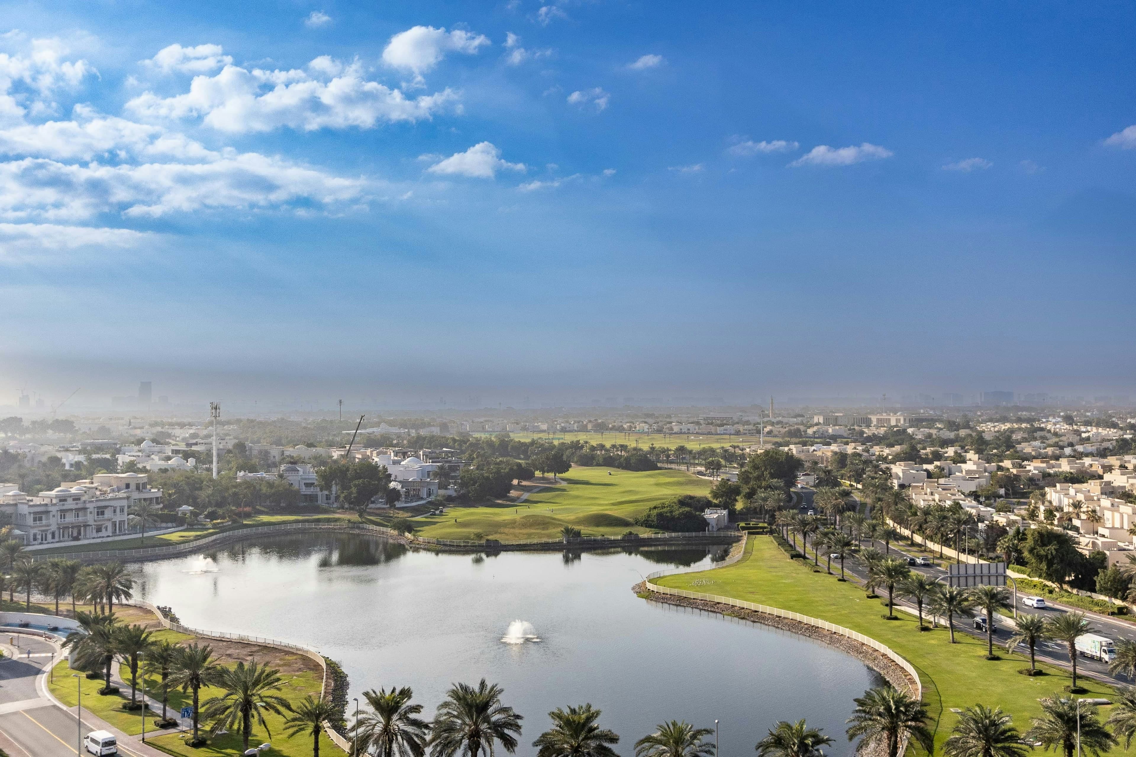 A lake with fountains in front of a fairway of the Montgomerie golf course in Emirates Hills, with large white villas on the left and palm-lined roads on the right