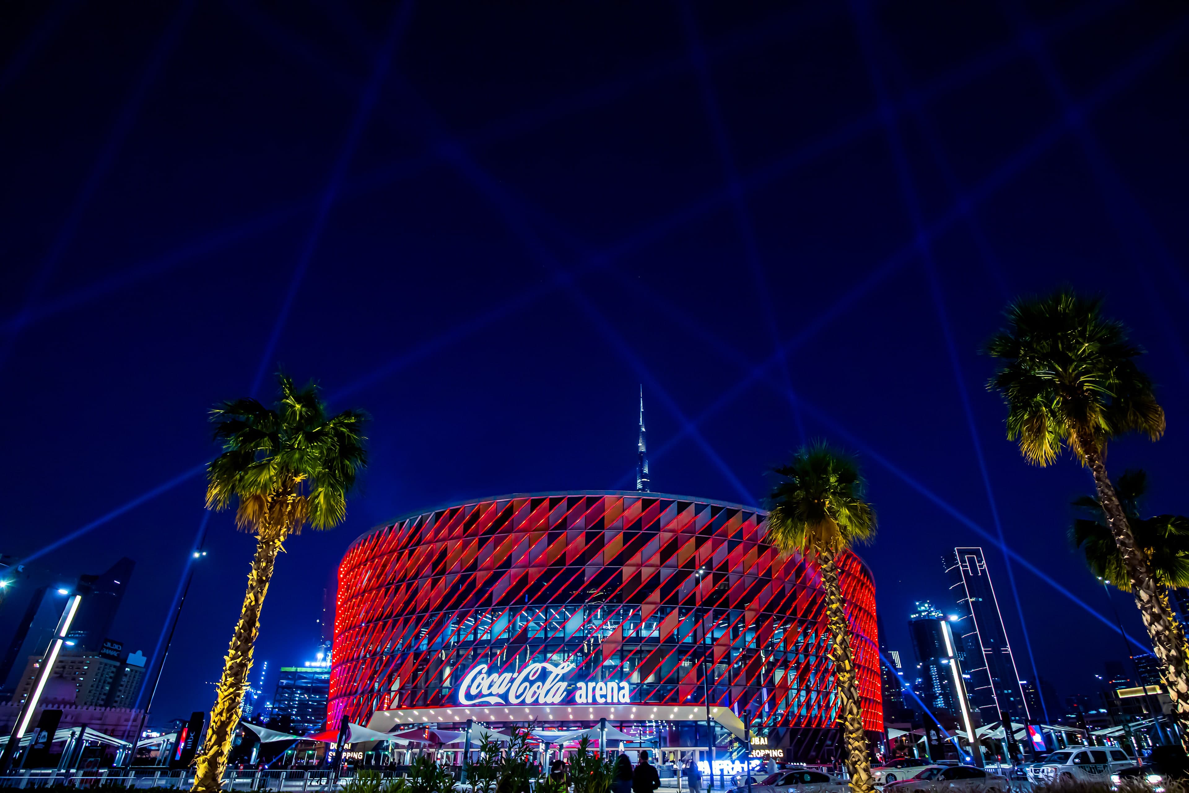 Coca-Cola Arena in City Walk lit up red at night, framed by palm trees, with the Burj Khalifa behind it
