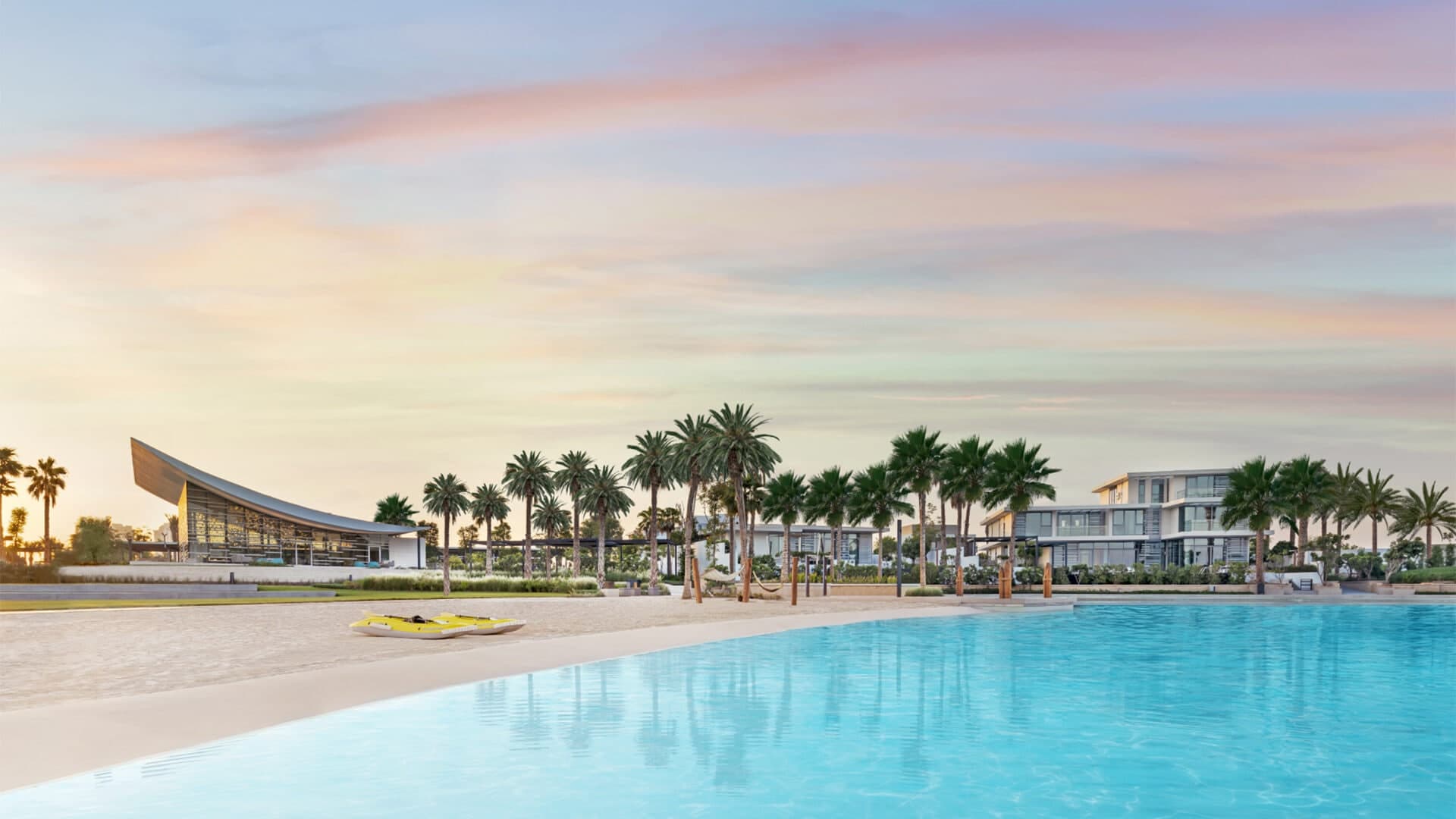 The sandy beach and turquoise water of Lagoon Al Ghaf in Tilal Al Ghaf, with palm trees, a curved clubhouse roof and lagoon-front homes behind