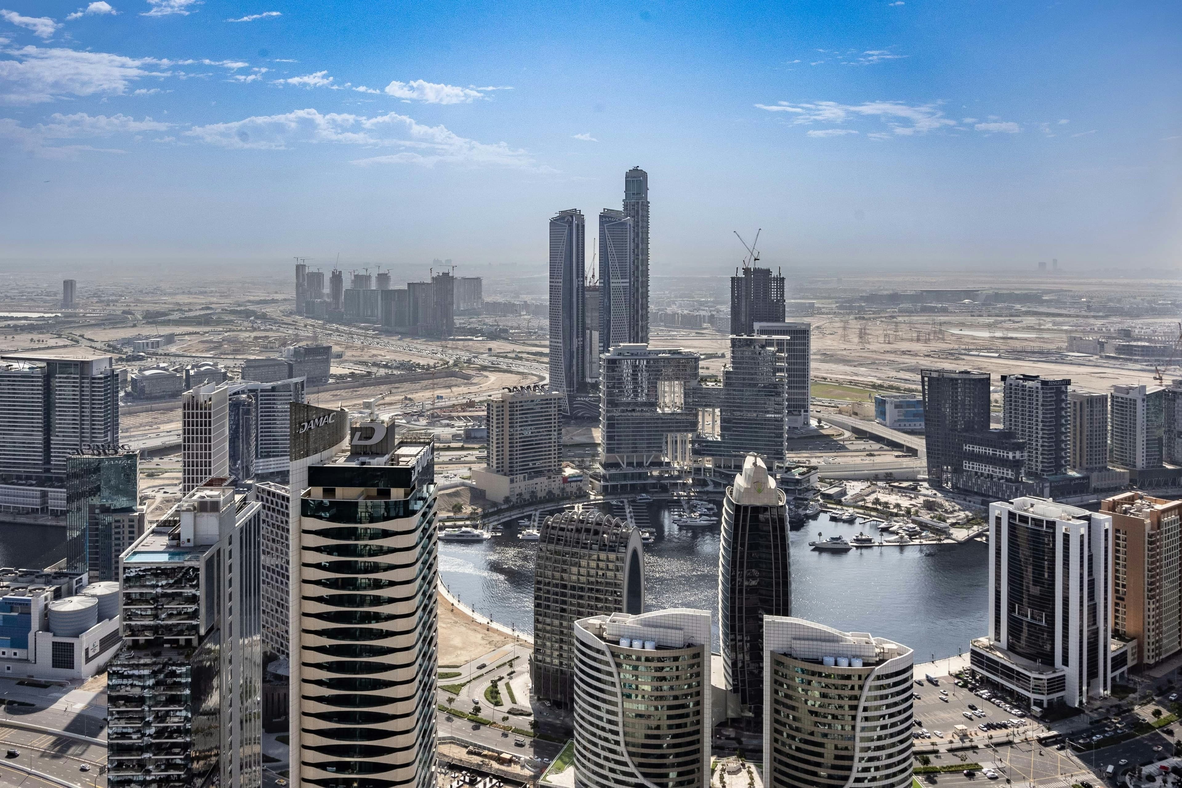 Business Bay from high above on a clear day, with towers around a canal basin where boats are moored and more towers under construction behind