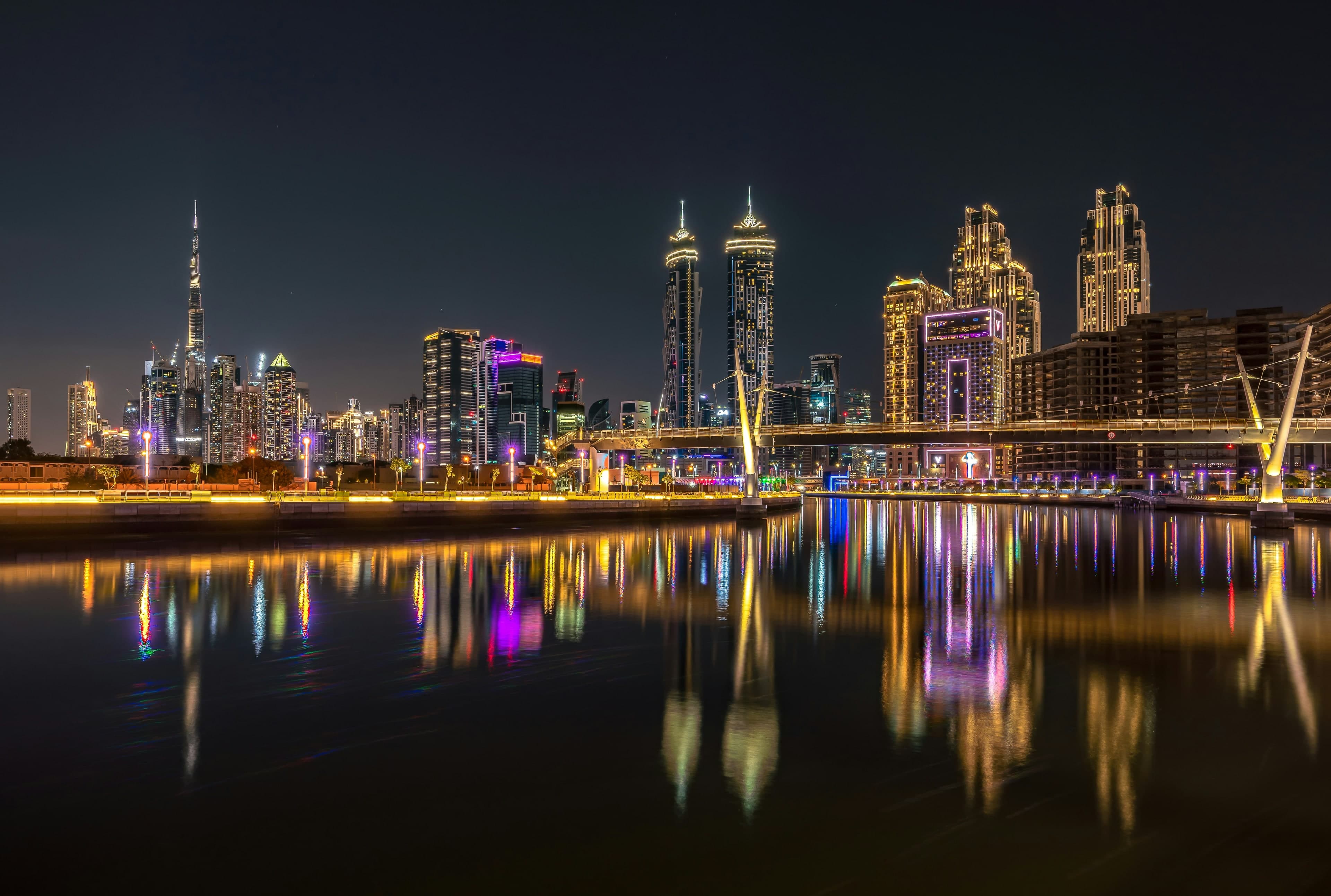 The Business Bay skyline at night reflected in the Dubai Water Canal, with a footbridge on the right and the Burj Khalifa on the left