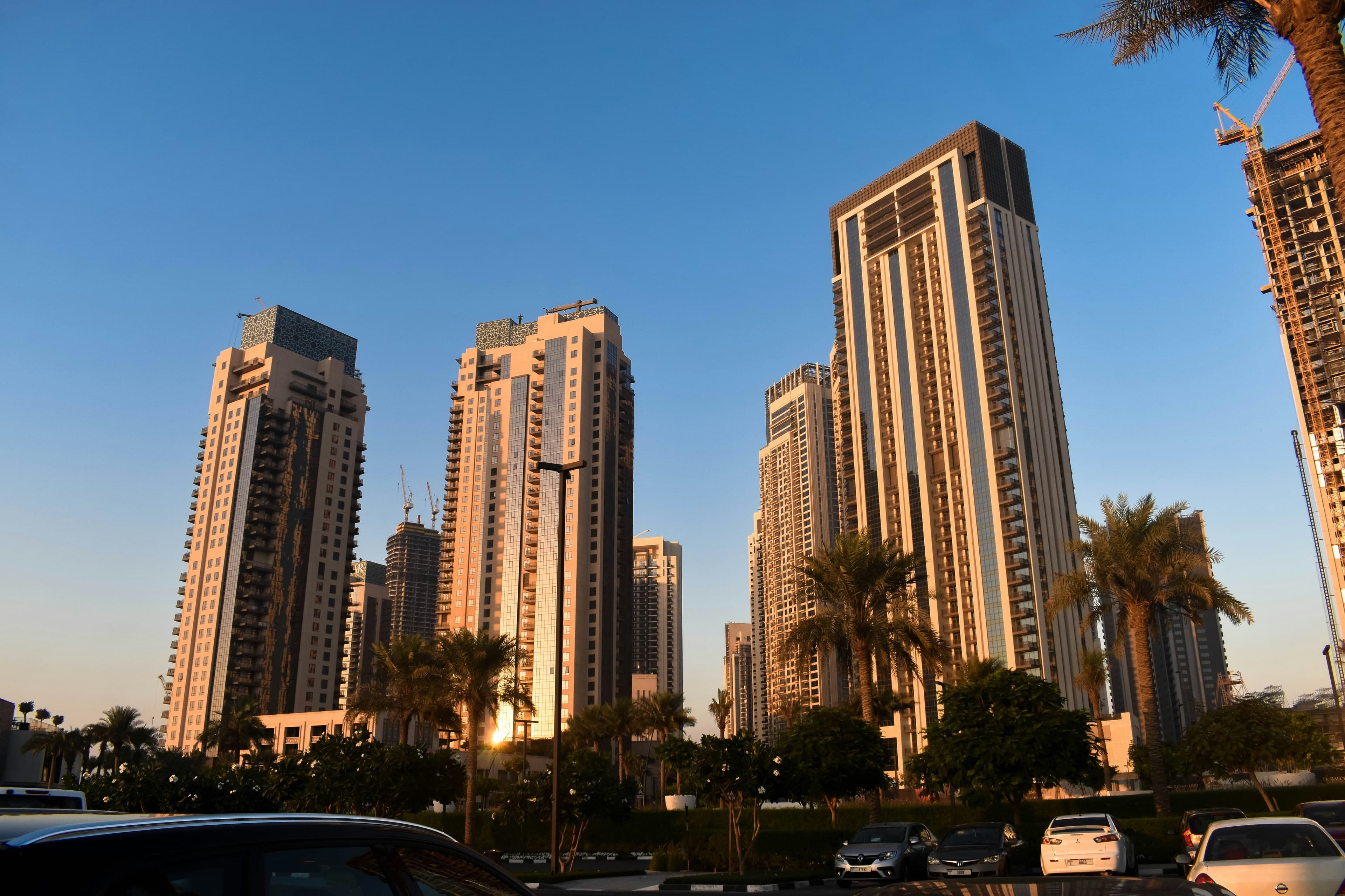 Residential towers of Dubai Creek Harbour in the late afternoon sun, with palm trees and a car park in front and a tower under construction on the right