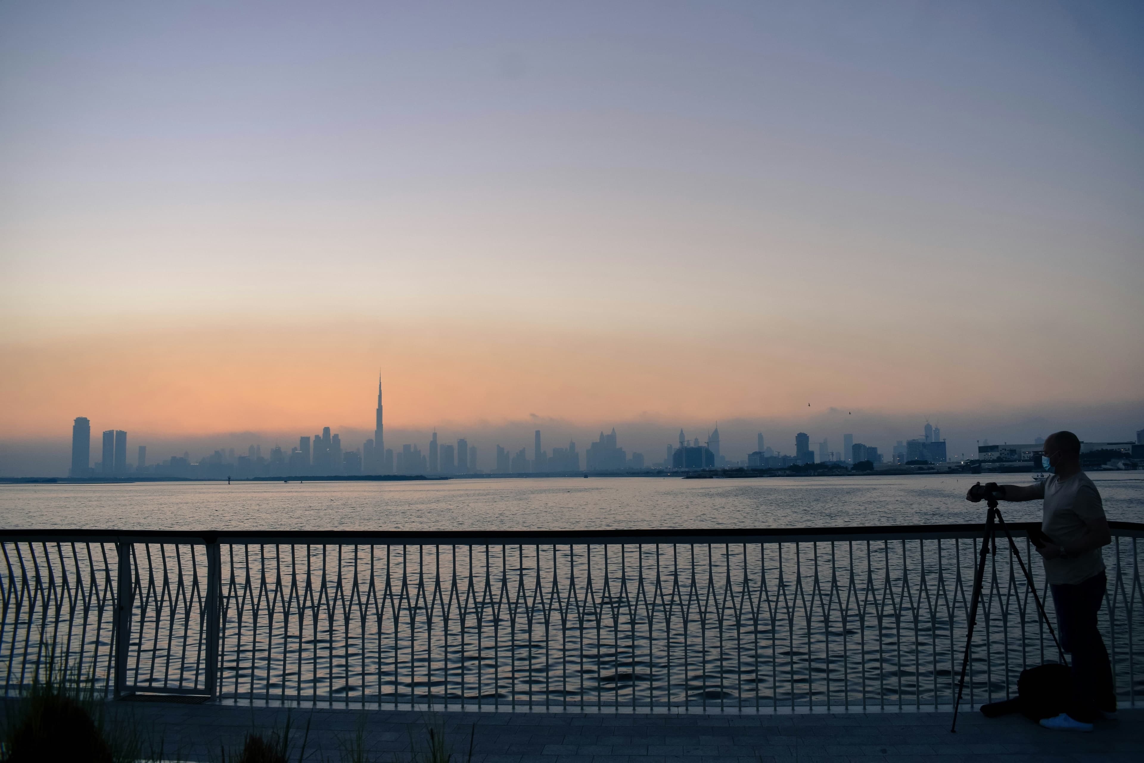 A photographer on the Dubai Creek Harbour promenade at dawn, looking across the water to the Downtown Dubai skyline