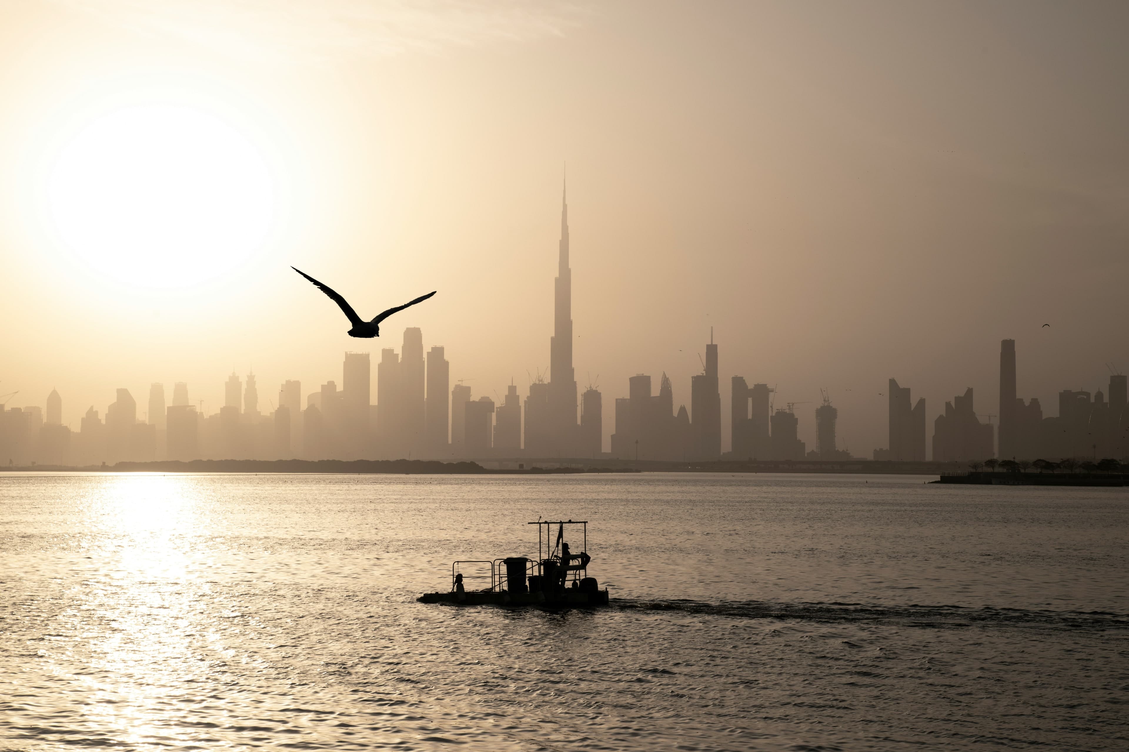 The Downtown Dubai skyline and the Burj Khalifa in a hazy sunset, seen across Dubai Creek from Dubai Creek Harbour, with a small boat and a gull in the foreground