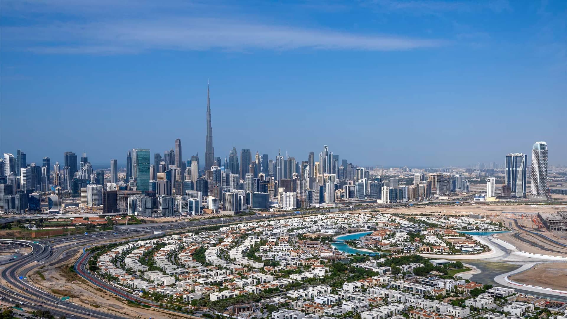 Aerial view of District One's curved rows of white villas beside Al Khail Road, with the Burj Khalifa and Business Bay towers in the distance