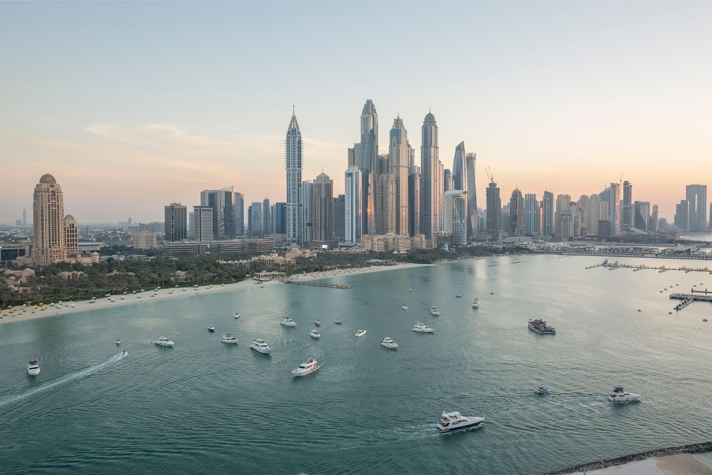 The Dubai Marina skyline seen across the water, with yachts in the bay and a sandy beach along the shore