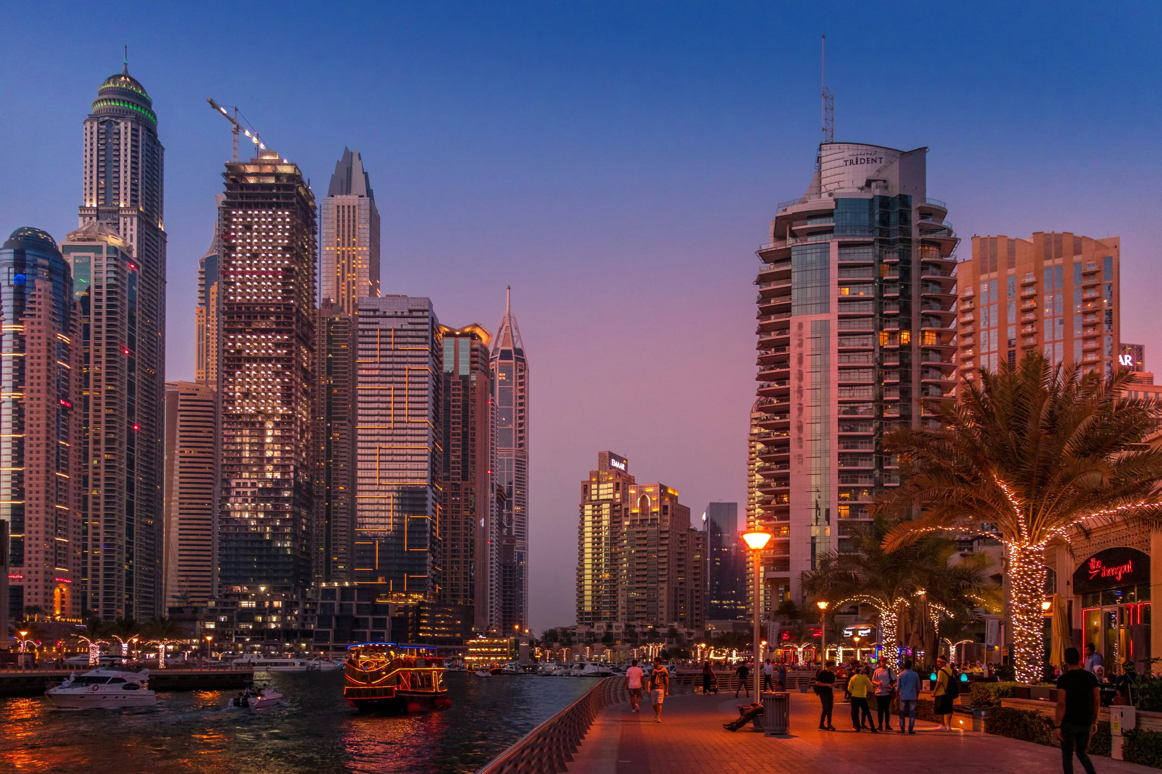 People walking along Marina Walk at dusk, with lit palm trees, a tour boat on the canal and towers rising behind